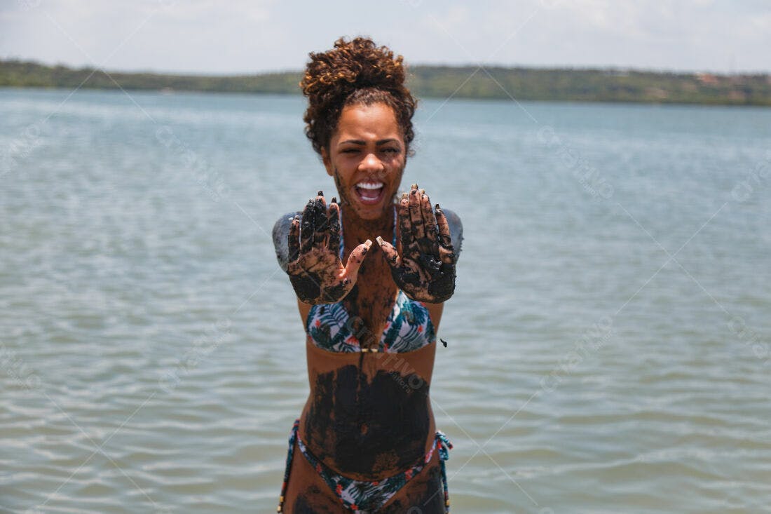 Mulher na praia com lama preta mostrando as mãos
