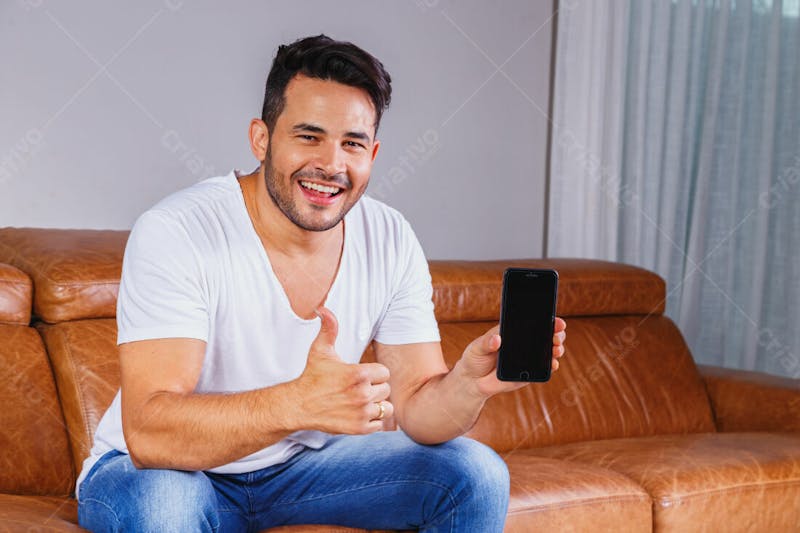 Homem jovem com celular na mão sentado no sofá da sala
