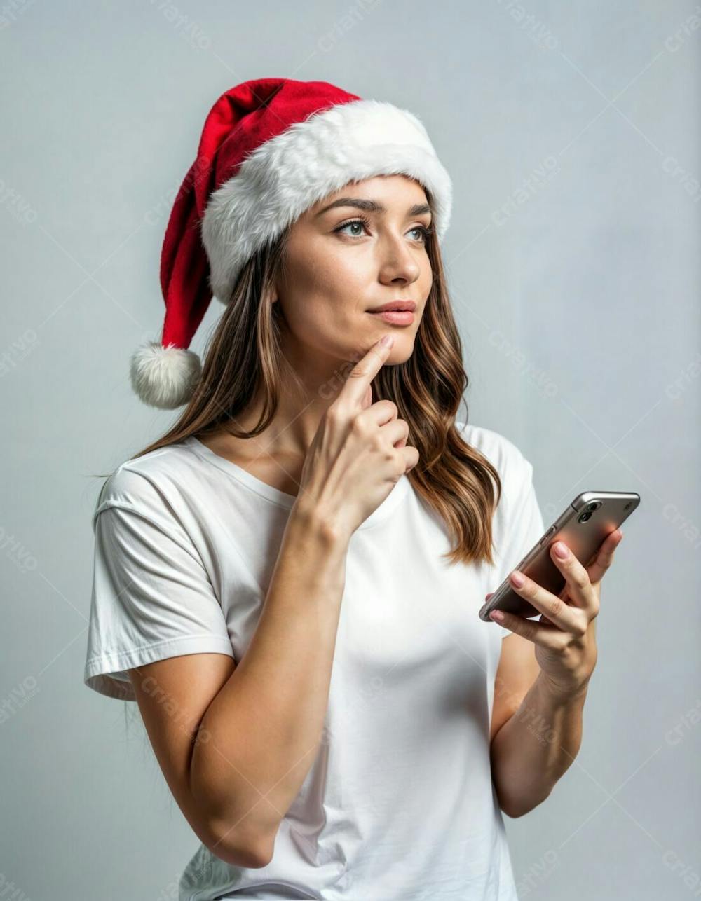 Linda Mulher Com Gorro De Papai Noel, De Camiseta Branca, Segura O Queixo E Pensa Sobre A Oferta On Line Do Celular, Desvia O Olhar E Contempla Algo, Isolado Sobre Um Fundo Cinza Claro Com Espaço Vazio