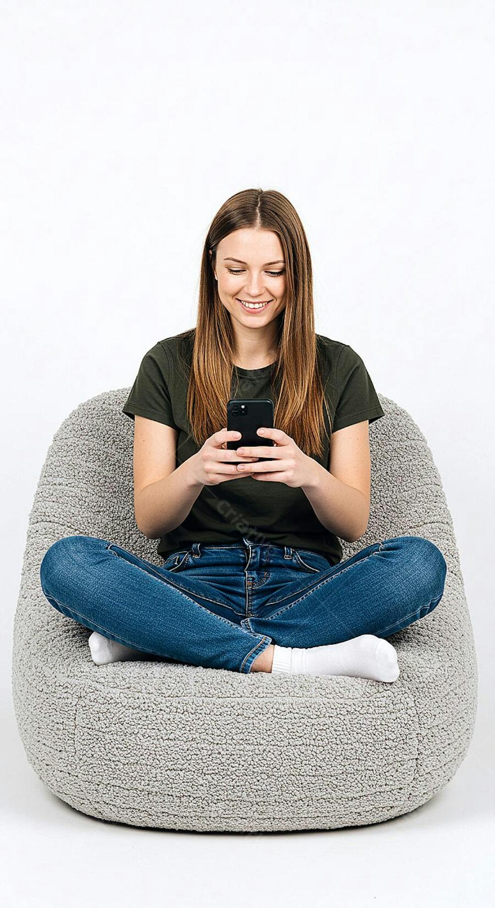 Uma jovem com cabelos longos e lisos está sentada de pernas cruzadas em uma cadeira confortável e grande absorta em seu smartphone