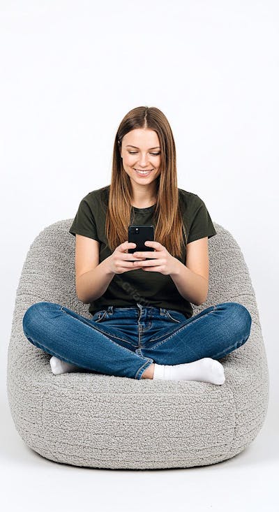 Uma jovem com cabelos longos e lisos está sentada de pernas cruzadas em uma cadeira confortável e grande absorta em seu smartphone
