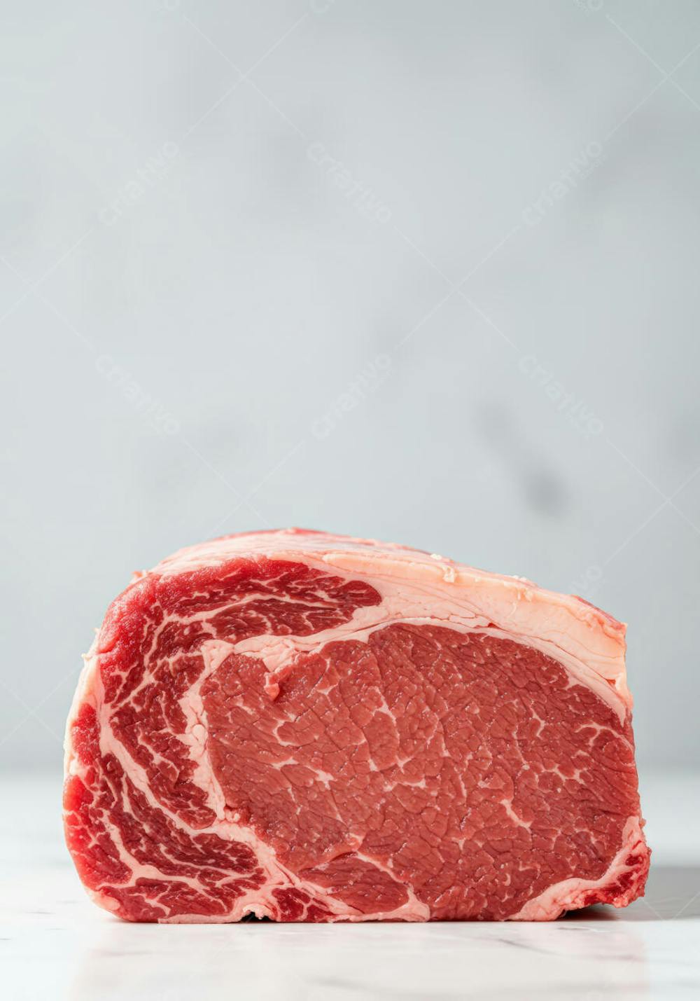 Imagem de um corte de Ribeye cru, com marmoreio visível, sobre uma superfície de mármore branco. As cores predominantes são o vermelho intenso da carne e o branco do mármore. A fotografia é simples e destaca a textura e a cor da carne.