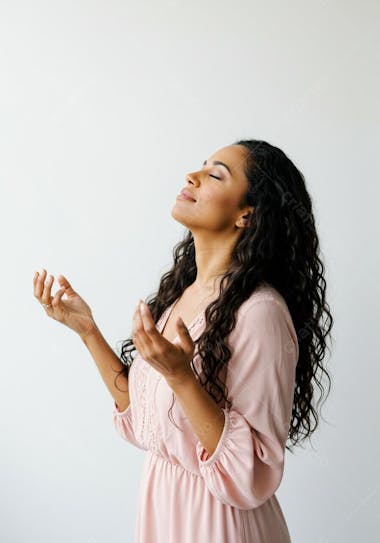 Photo of a brown-skinned woman with long, curly hair wearing a light pink dress.  She has her eyes closed and hands raised in a meditative or prayerful pose against a white background. The mood is serene and calm with soft colors and natural lighting.