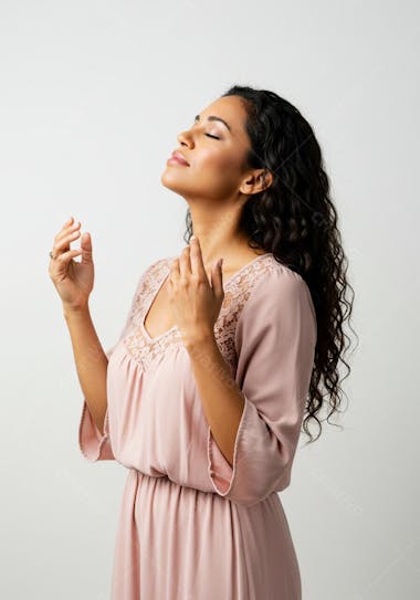 Image of a woman with her eyes closed, dark curly hair and brown skin, wearing a light pink dress with lace details. The atmosphere is calm and serene, with a neutral white background. The composition focuses on the woman, highlighting her peaceful expression.