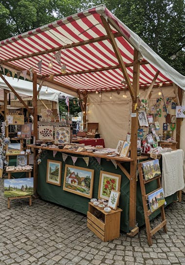 Wooden stall with red and white striped awning, selling paintings of houses and landscapes in shades of green, blue, and brown.  Various handmade crafts and decorative objects are also displayed. The atmosphere is cozy and idyllic.