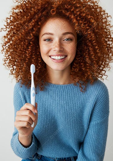 Close-up shot of a young Caucasian woman with curly redhead hair and freckles smiling at the camera while holding a toothbrush. Predominant colors are blue, white and copper-red tones. The mood is cheerful and positive.