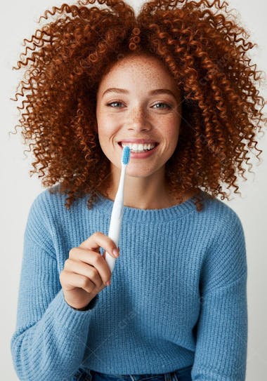 Close-up shot of a young woman with curly red hair and freckles, wearing a light blue sweater, holding a toothbrush and smiling at the camera. The image features pastel tones and a cheerful, healthy atmosphere.
