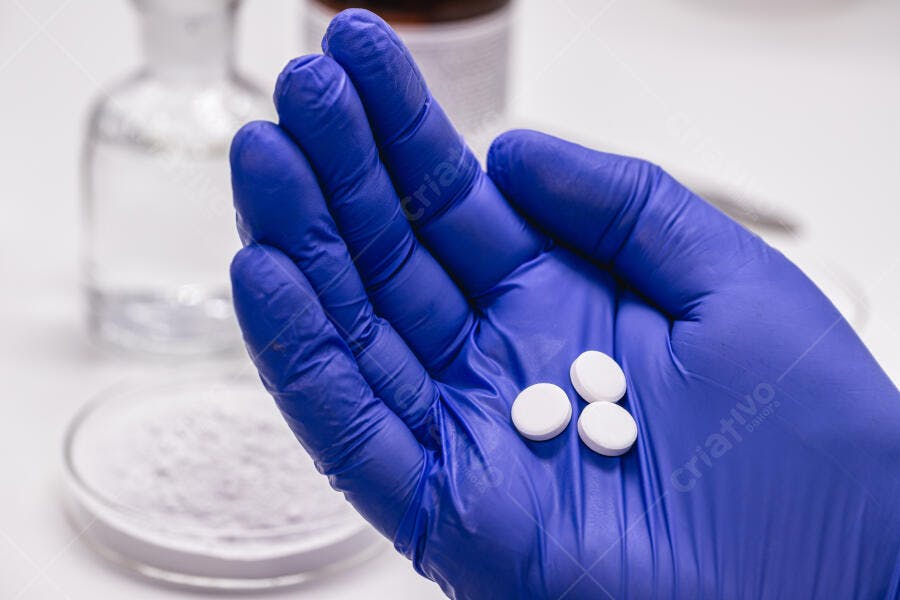 Mão com luva azul segurando três comprimidos brancos em laboratório