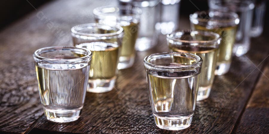 Doses de bebidas alcoólicas transparentes e douradas em mesa de madeira rústica