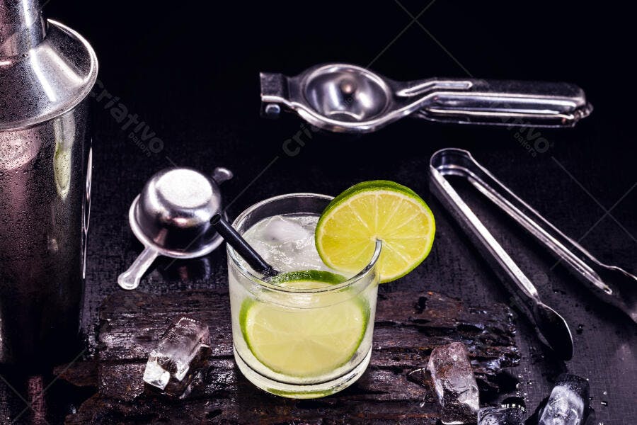 Coquetel de caipirinha com lima e gelo em bancada escura com utensílios de bar