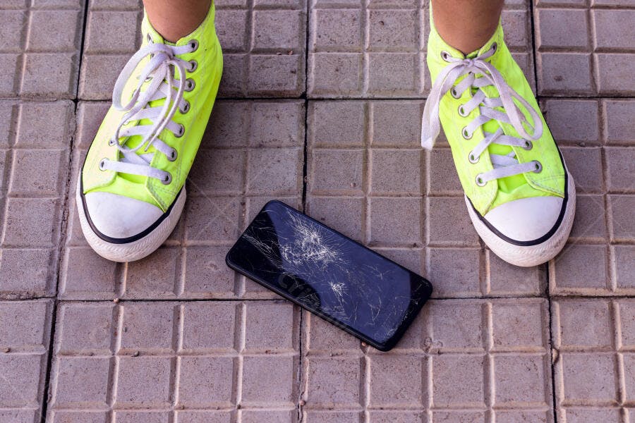 Celular com tela quebrada no chão perto de tênis amarelo neon