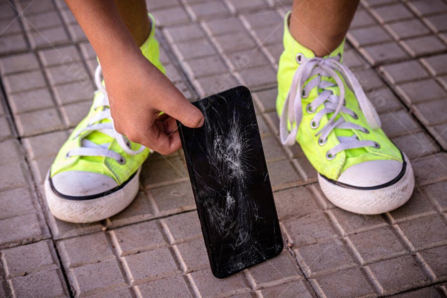 Criança com tênis amarelos segura celular tela quebrada na calçada