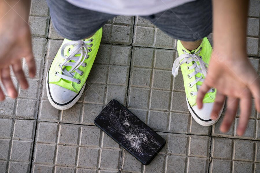 Pessoa de tênis neon olhando celular com tela quebrada no piso de azulejos