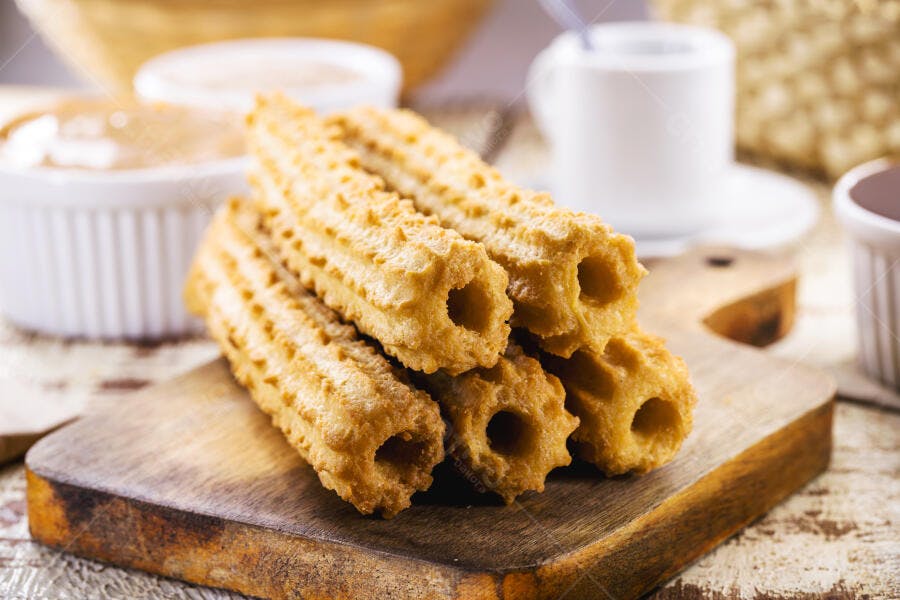 Churros dourados crocantes em tábua de madeira com molhos e café no fundo