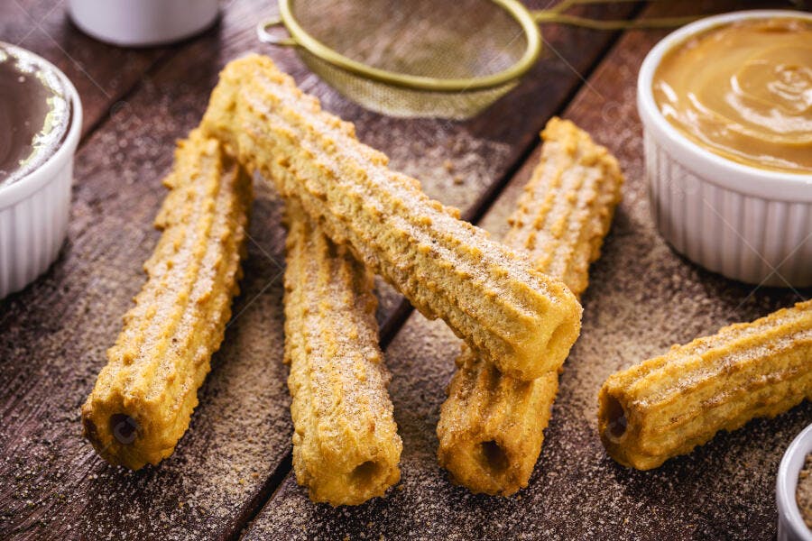 Churros crocantes com açúcar e canela servidos com doce de leite e chocolate em mesa de madeira
