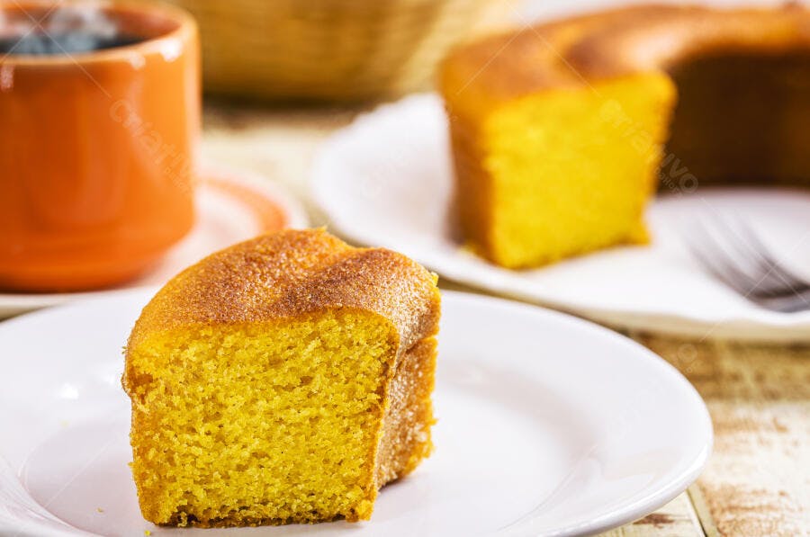 Fatia de bolo de milho amarelo em prato branco com caneca laranja na mesa de madeira