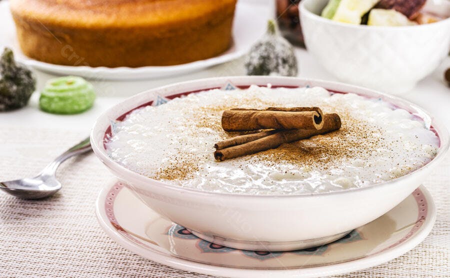 Arroz doce cremoso com canela em tigela branca e bolo de festa ao fundo