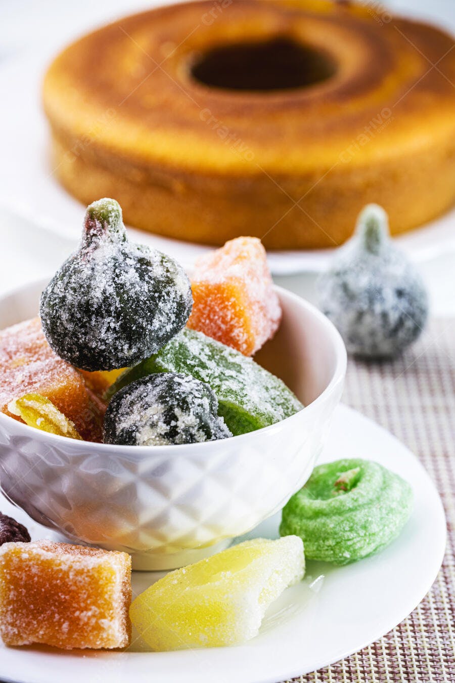 Doces cristalizados coloridos em tigela branca com bolo de fubá no fundo