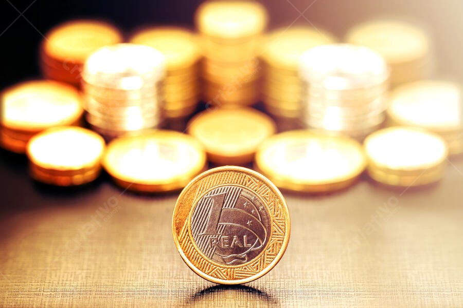 Moeda de um real brasileira em pé com pilhas de moedas douradas no fundo