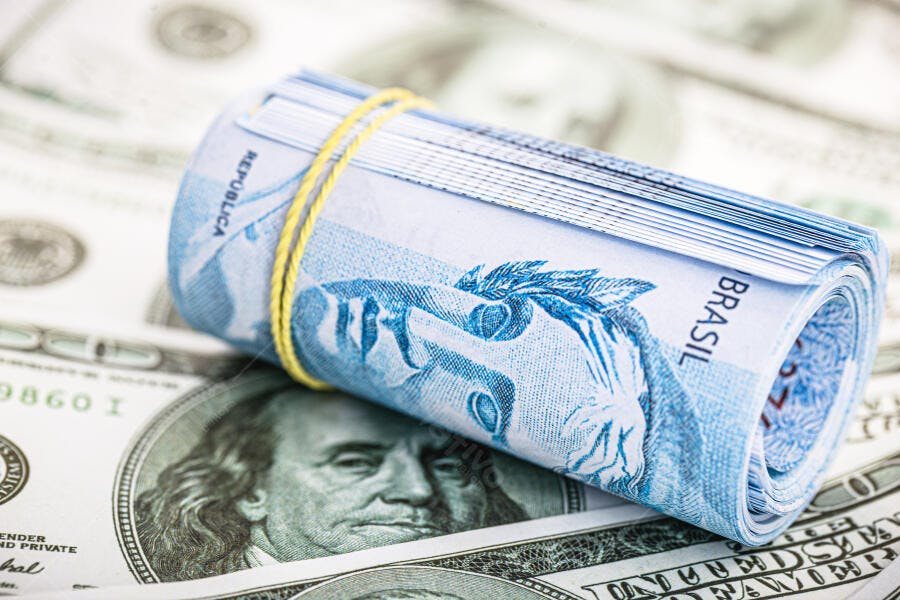 Cédulas de reais azuis enroladas com elástico amarelo sobre notas de dólar
