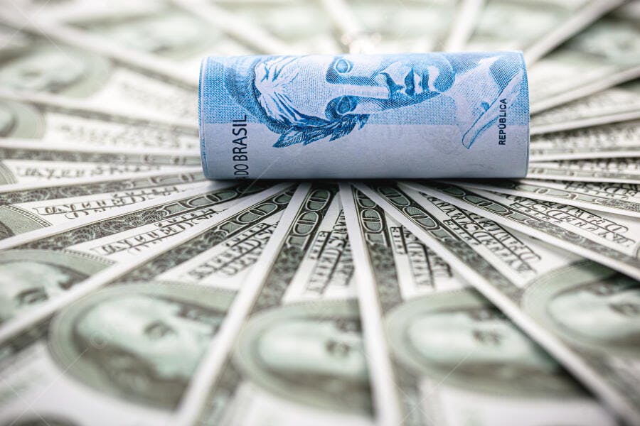 Nota de 100 Reais azul enrolada sobre notas de Dólar americano espalhadas