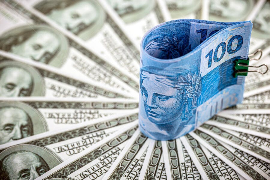 Cédula de 100 Reais azul em destaque sobre dólares americanos em leque