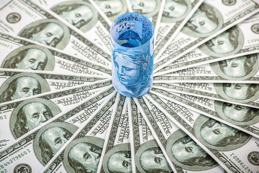 Cédula de 100 Reais azul rodeada por dólares americanos verdes em composição financeira