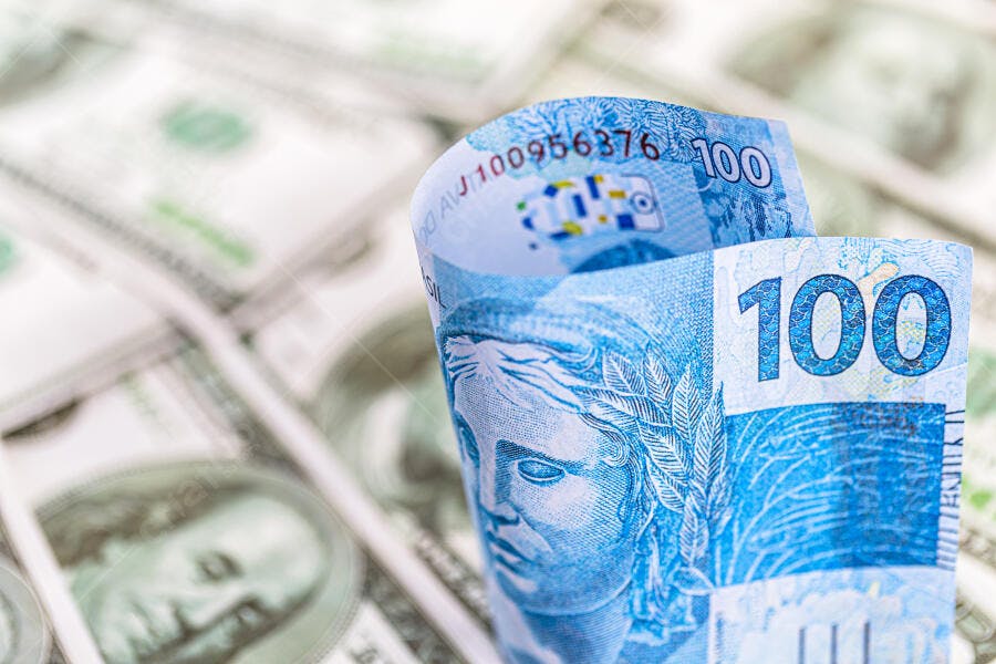 Cédulas de 100 Reais em destaque com Dólares americanos no fundo