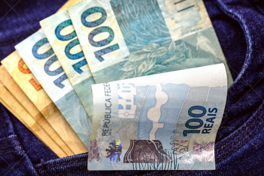 Cédulas de 100 e 50 Reais azuis e amarelas no bolso de uma calça jeans azul