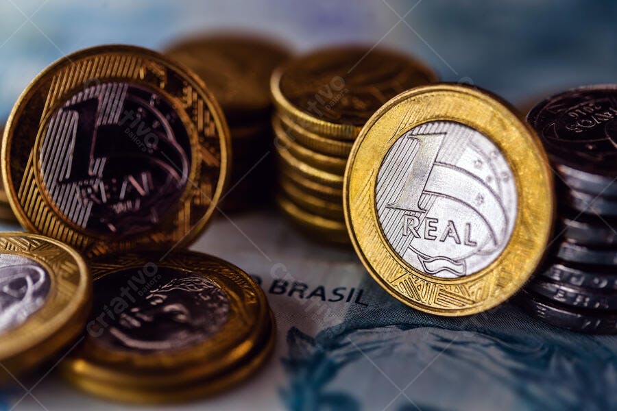 Moedas de um Real douradas e prateadas empilhadas sobre nota de dinheiro do Brasil