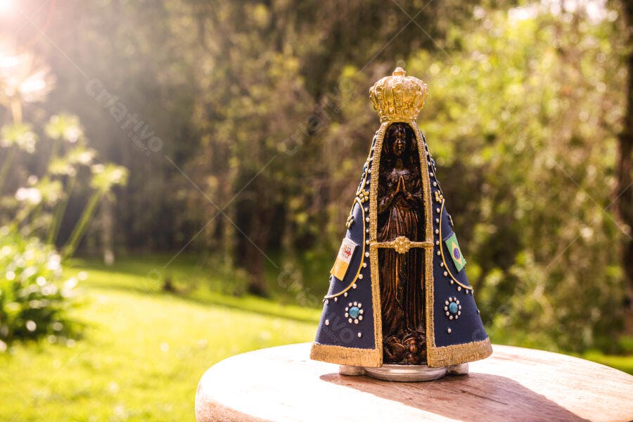 Estátua de Nossa Senhora Aparecida em jardim ensolarado com bandeira do Brasil