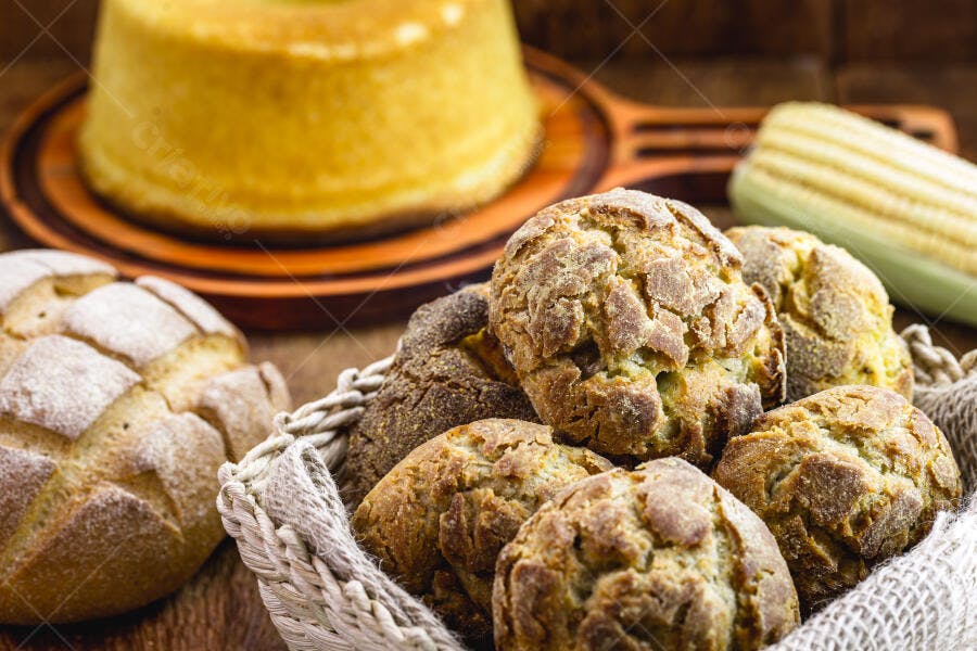Pães de milho em cesta rústica com bolo de fubá e espiga de milho na mesa de madeira