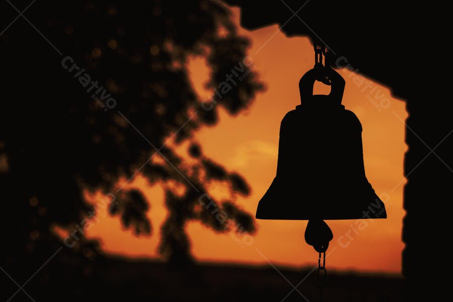 Sino em silhueta contra pôr do sol laranja com ramos de árvore