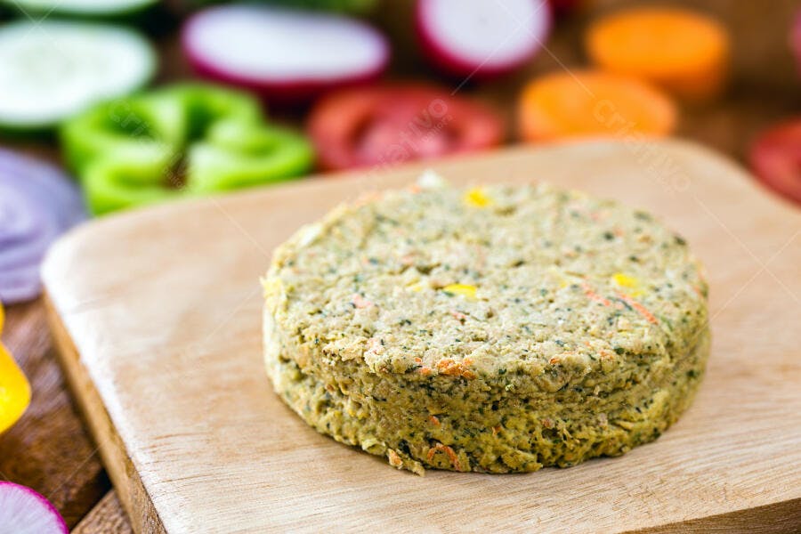 Hambúrguer vegetal cru em tábua de madeira com legumes frescos ao fundo