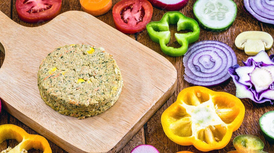 Hambúrguer vegetariano verde com milho em tábua de madeira rodeado por legumes vibrantes