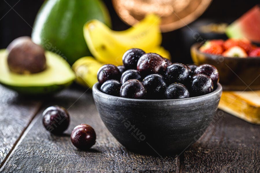 Jabuticabas frescas e roxas em tigela escura com abacate e banana ao fundo