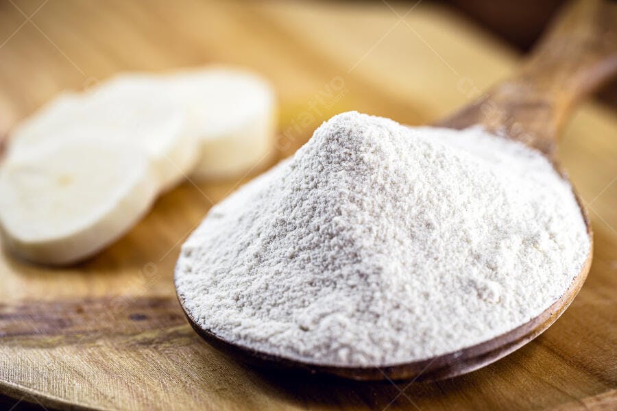 Farinha de mandioca branca em colher de pau com fatias de raiz na tábua de madeira