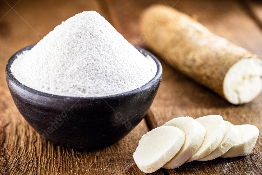 Farinha de tapioca branca em tigela com mandioca crua e fatiada na madeira
