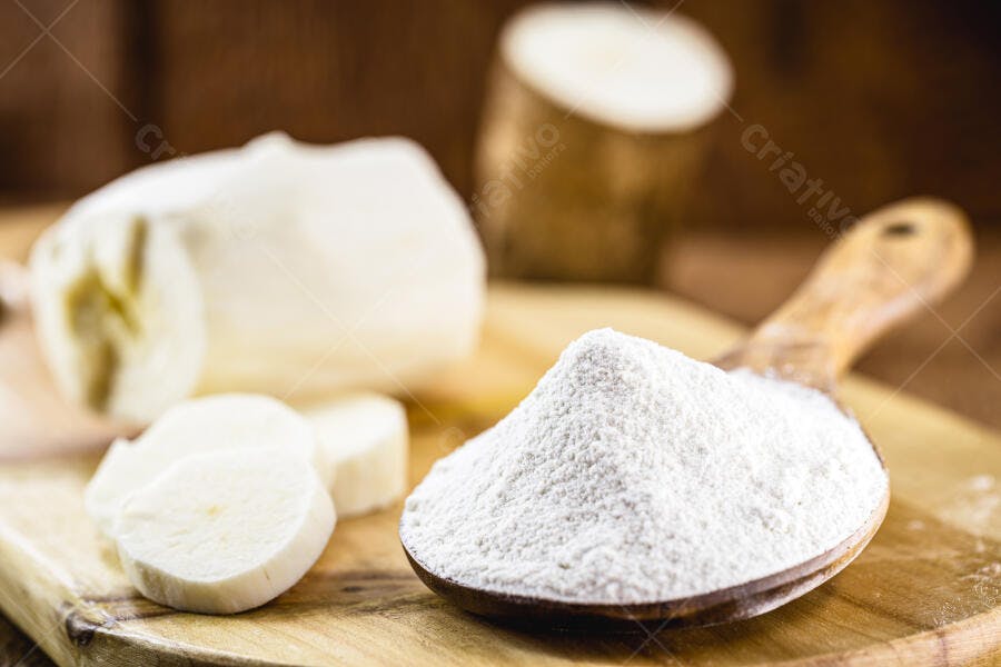 Farinha de mandioca branca na colher de pau com raízes cruas na tábua de madeira