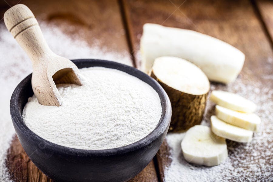 Farinha de mandioca em tigela preta com colher de pau e pedaços de mandioca crua