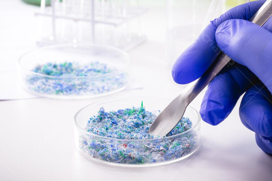 Mão de cientista com luva azul manuseando microplásticos coloridos em laboratório