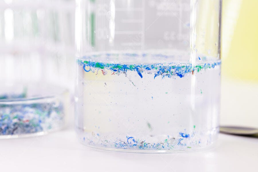 Microplásticos azuis e verdes flutuando na água em béquer de laboratório