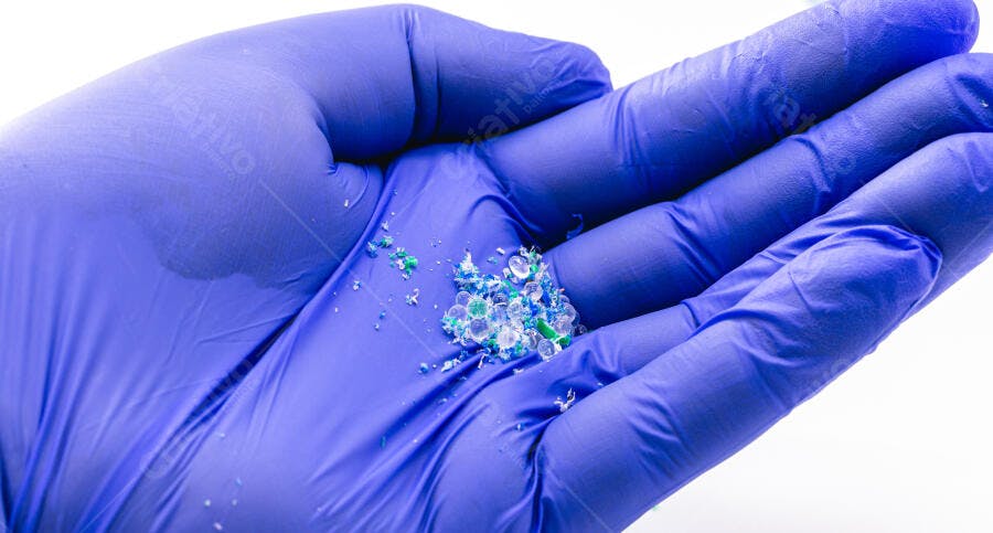 Mão enluvada azul segurando microplásticos coloridos granulados em fundo branco
