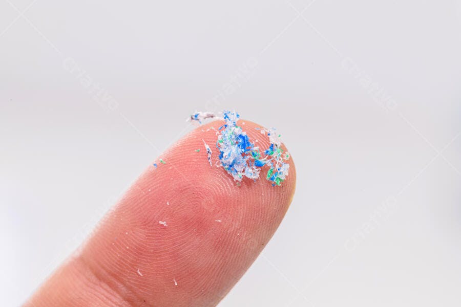 Dedo humano segurando microplásticos azuis e verdes em fundo branco