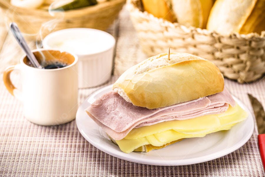 Pão francês com presunto e queijo, café quente fumegante e cesta de pães