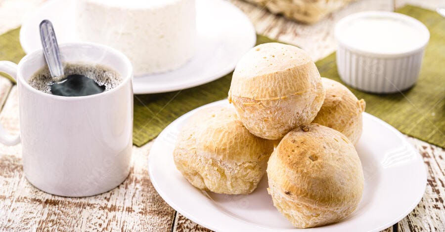 Pão de queijo dourado, café em caneca branca, queijo fresco e iogurte para café da manhã
