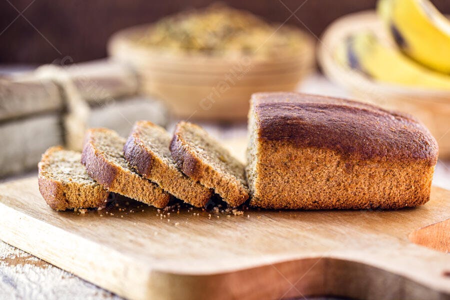 Pão de banana caseiro fatiado sobre tábua de madeira com frutas no fundo