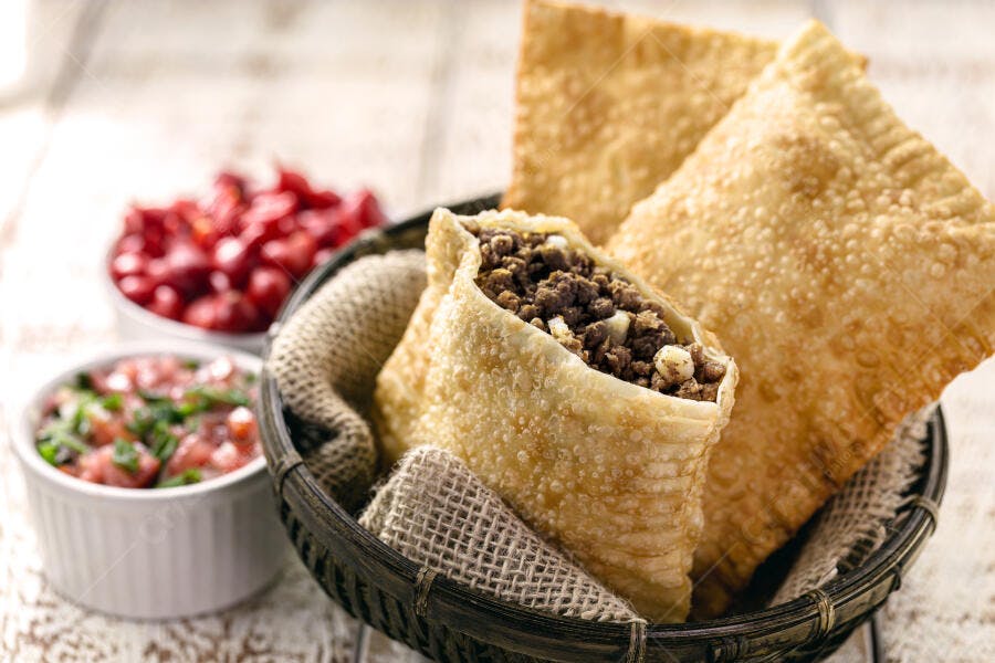 Pastéis fritos com recheio de carne moída em cesta com molhos sobre mesa rústica