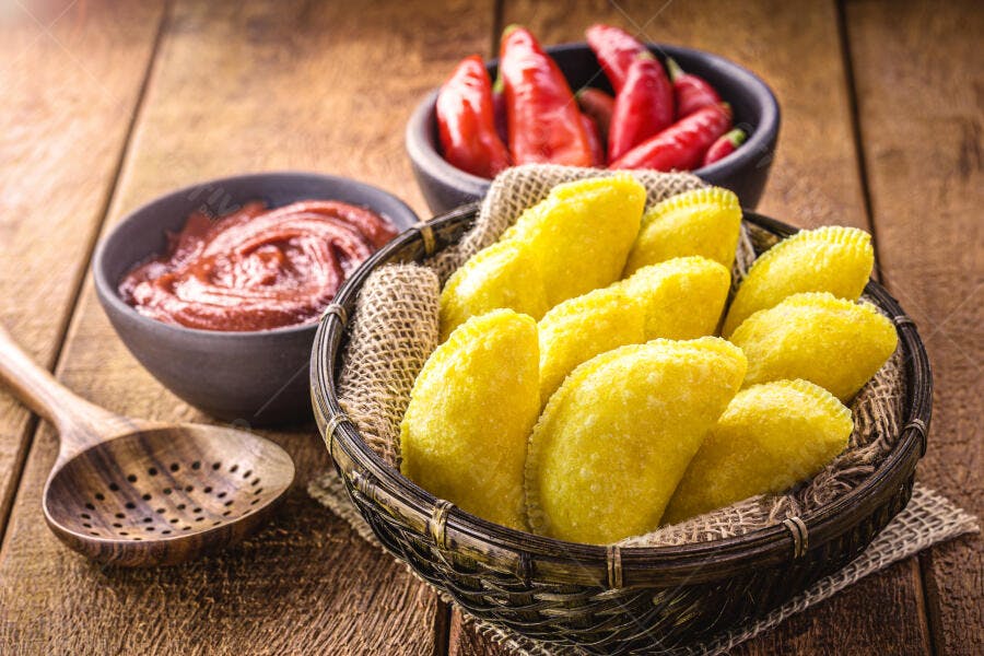 Pastéis fritos amarelos em cesta rústica com molho e pimentas vermelhas na mesa