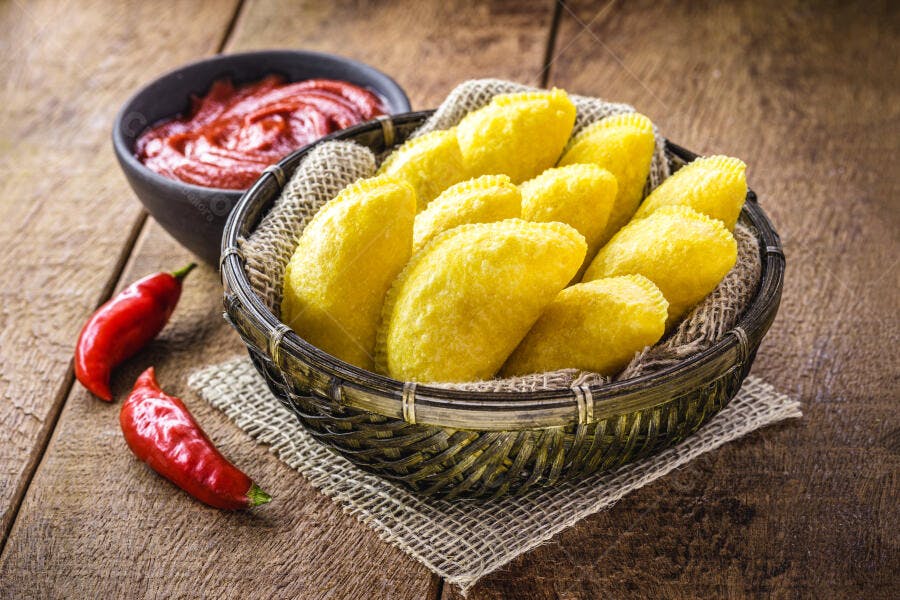 Pastéis fritos dourados em cesta com molho vermelho e pimentas na mesa de madeira